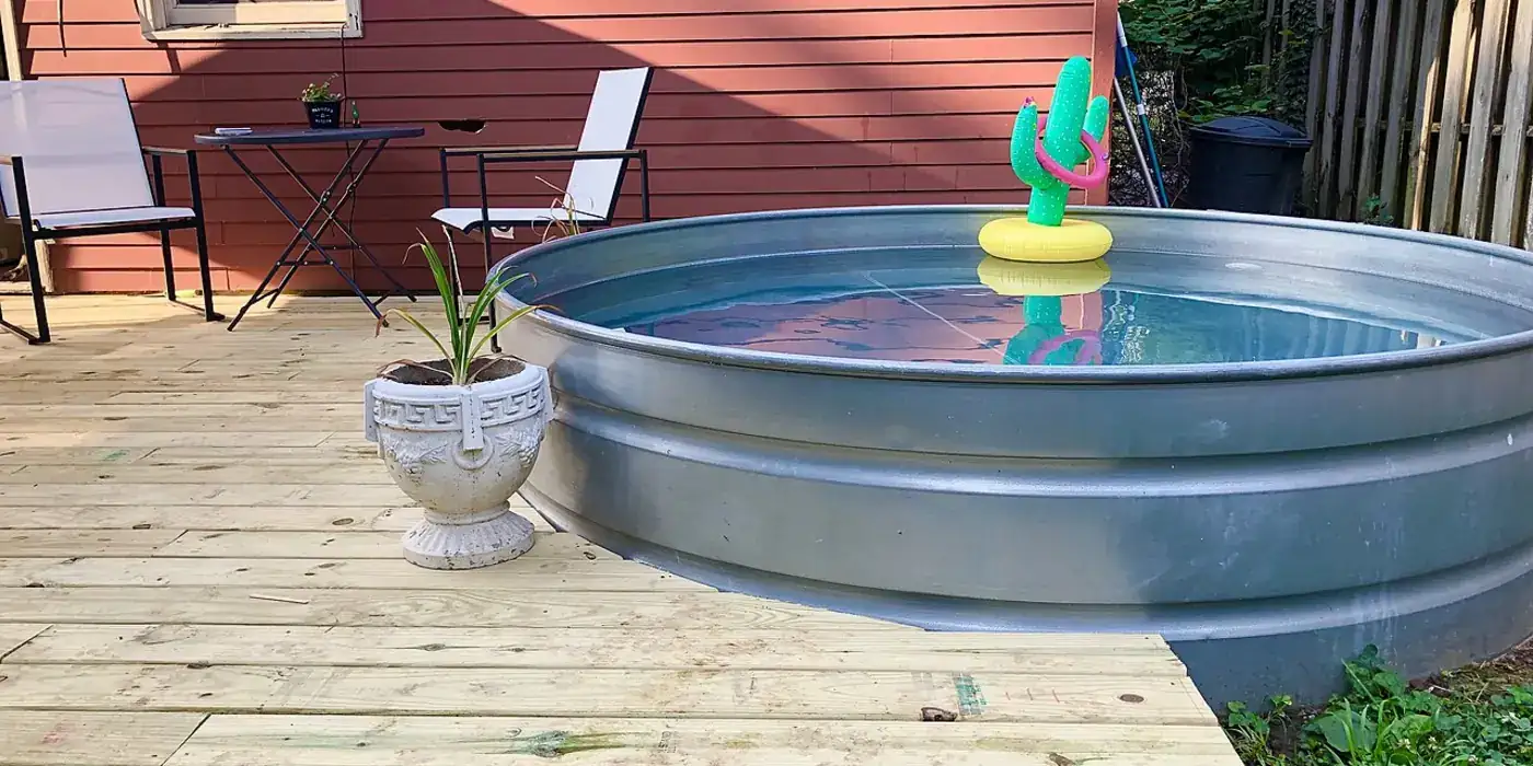 A round galvanised steel stock tank filled with water, standing on a new wooden deck in a small back garden next to two patio chairs.
