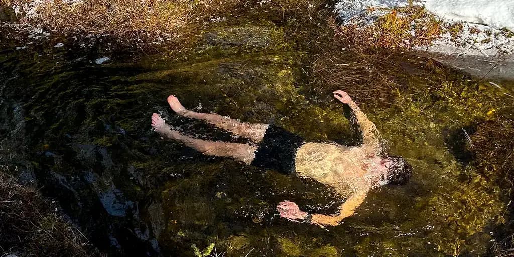 A person lies back in a shallow, weed-lined natural rock pool, submerged to the shoulders, with snow banked up on the rocks behind them.