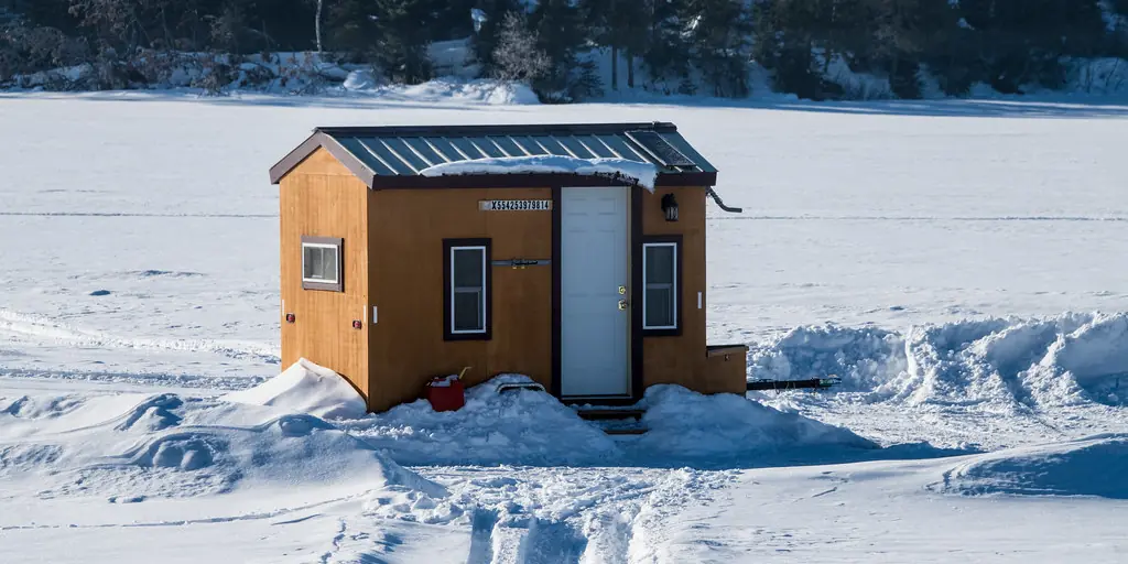 A single small ice-fishing shack sits alone on a wide frozen lake, with snowmobile tracks curving away across the snow towards a treeline.