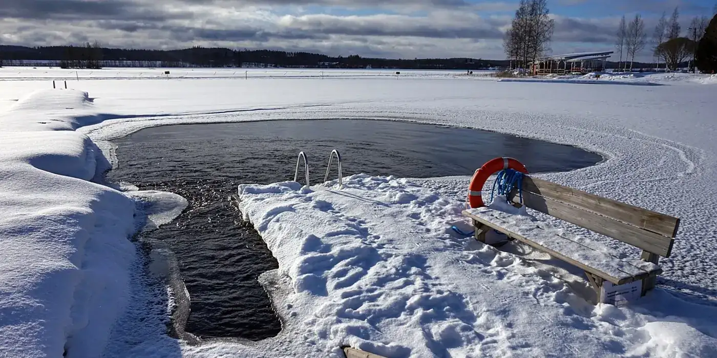 A rectangular hole cut into the ice of a frozen lake, with a metal ladder dropping into the black water, a bench and a lifebuoy on the snow beside it.