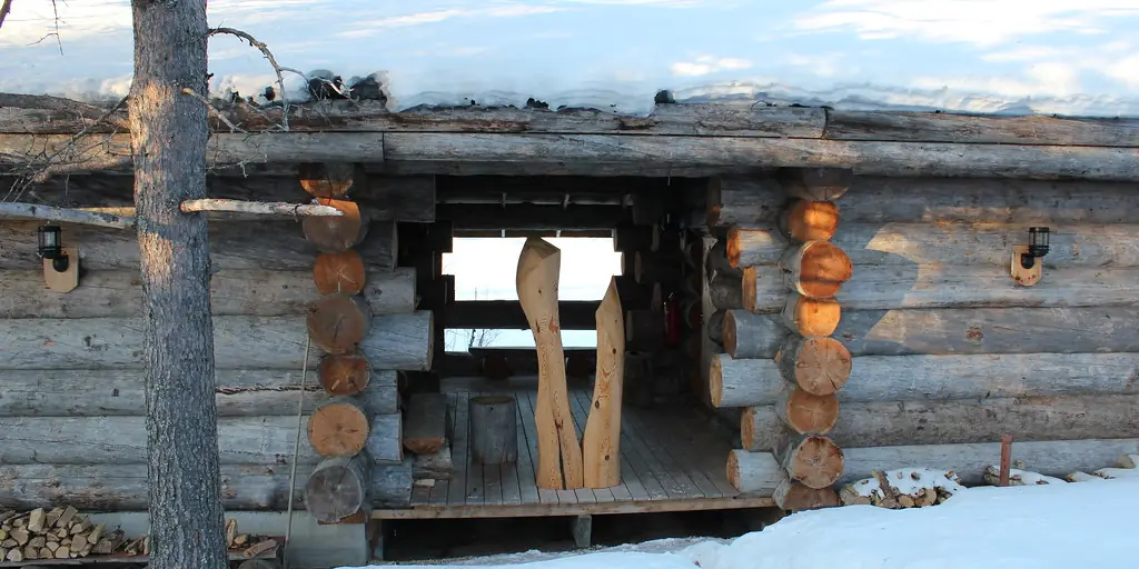 A small log-built sauna hut half buried in deep snow in a Finnish forest, with a wooden paddle propped against the doorway.
