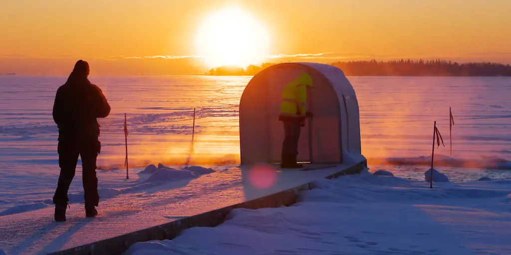 Two figures stand on a frozen lake at sunset beside a small curved changing hut, next to a swimming hole kept open in the ice.