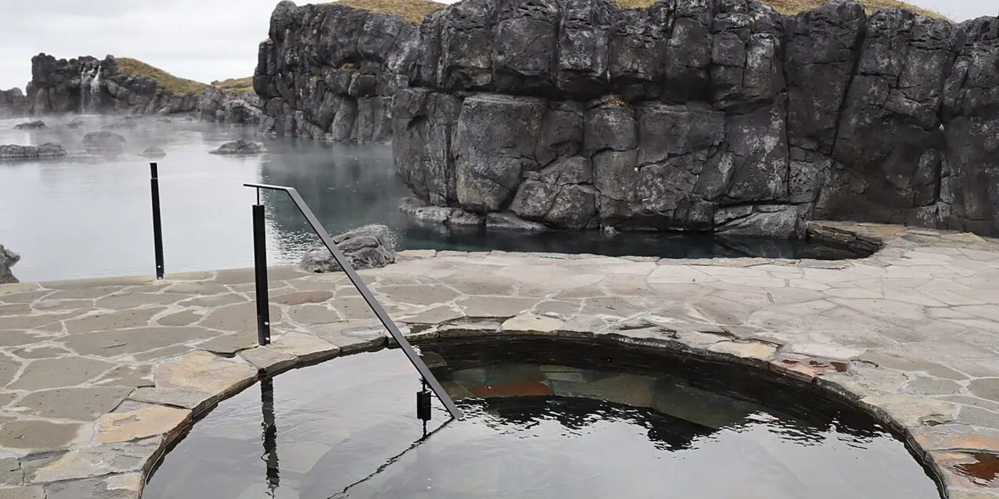 A circular stone-edged cold plunge pool set into a paved terrace beside a lava rock wall, with steam drifting off warmer water behind it.