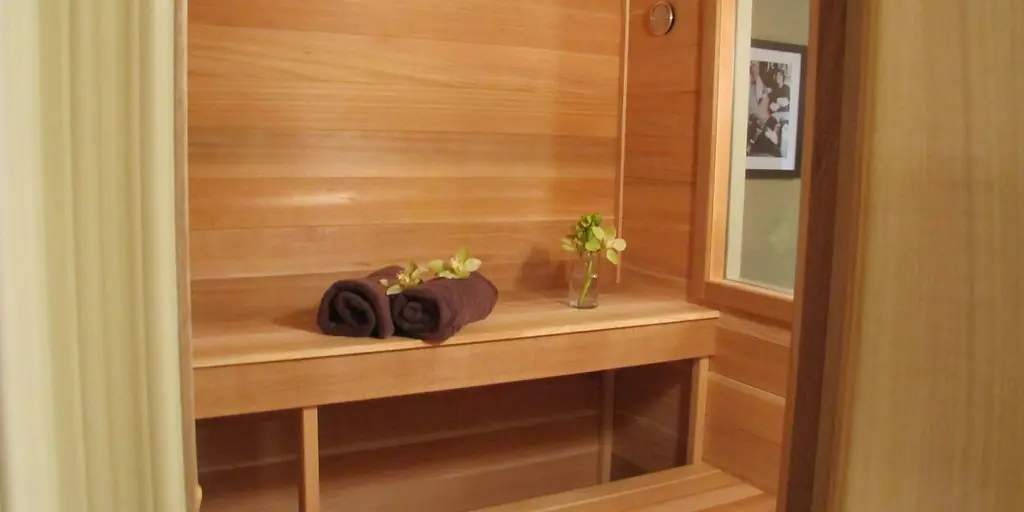 The inside of a small cedar-lined sauna cabin, with a slatted bench, rolled towels and a glass door letting in daylight.