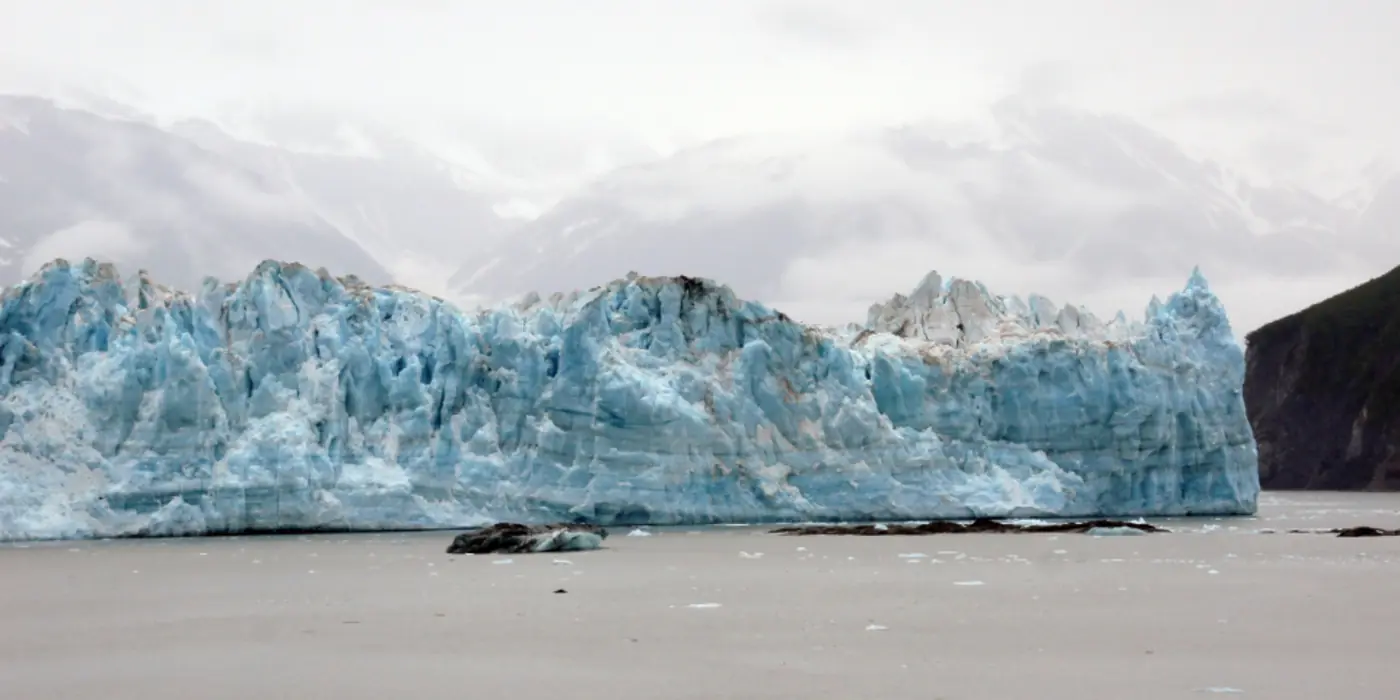 The blue-white face of a glacier meeting flat grey water under an overcast sky, with a dark shore in the foreground.