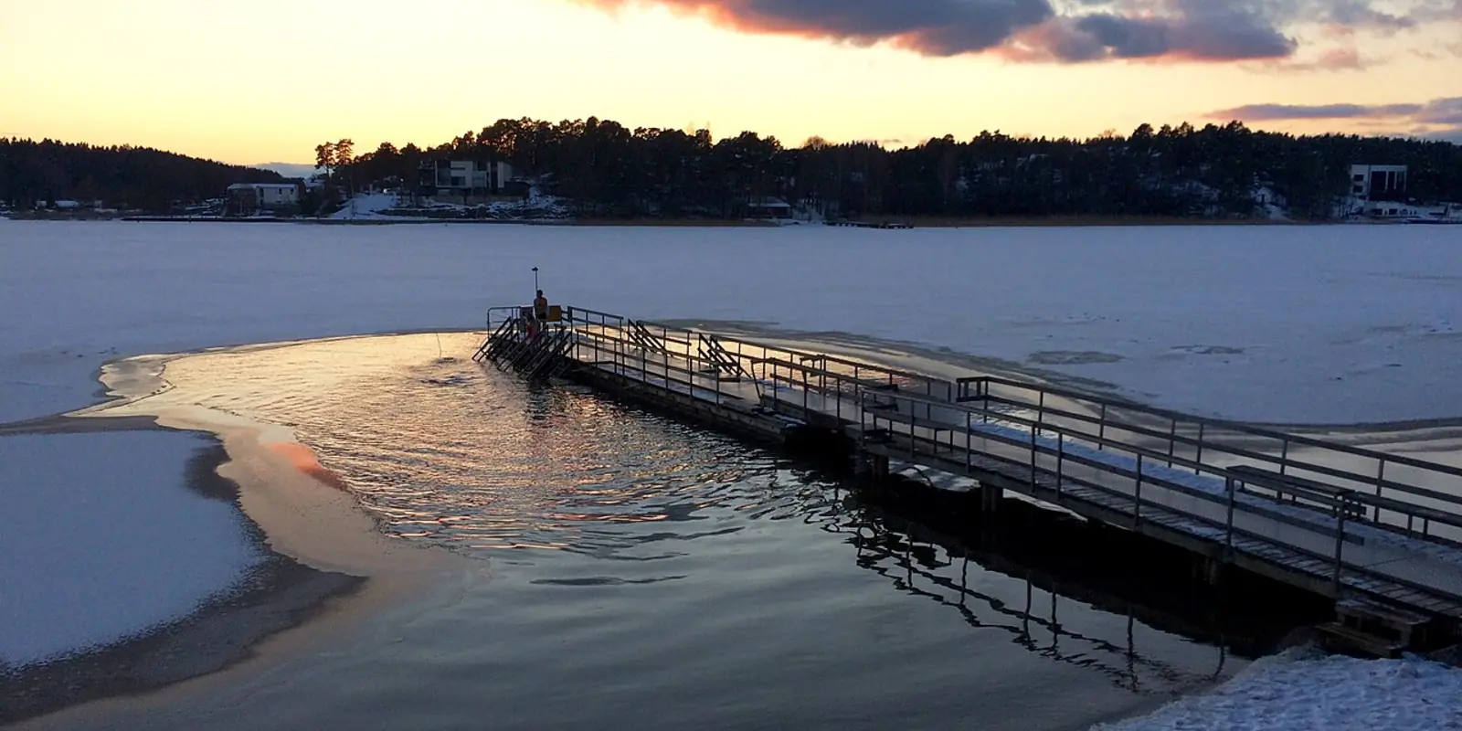 A wooden jetty runs out across a frozen bay at dusk. A channel has been cut through the ice at the end of it, and the open water reflects an orange sunset.