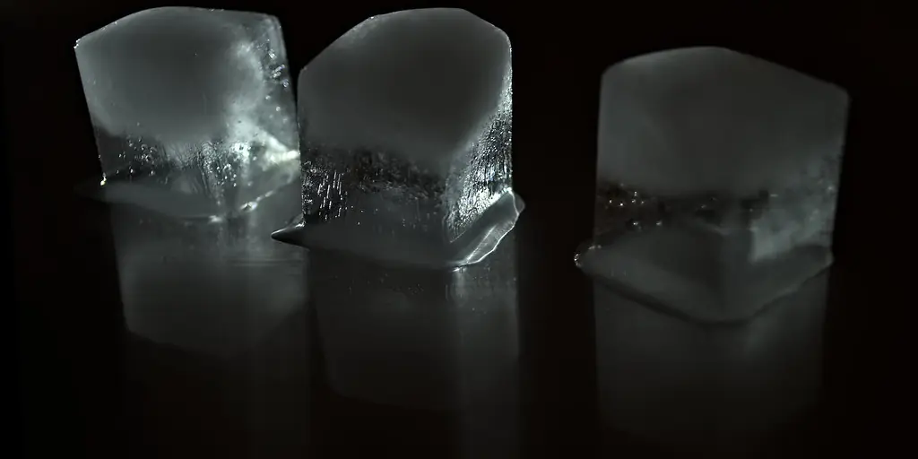 Three melting ice cubes sitting on a wet black surface against a dark background, lit so their edges glow.