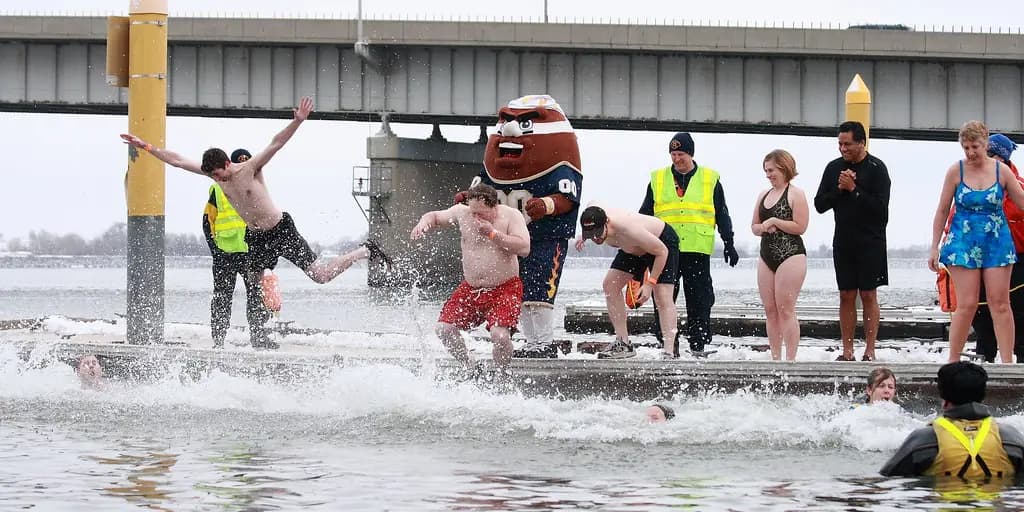 A line of people in swimsuits runs off a beach into grey winter surf at a charity polar plunge, throwing up white water around their legs.