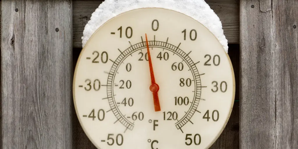 A round outdoor dial thermometer screwed to a weathered fence, capped with snow, its needle pointing well below freezing.