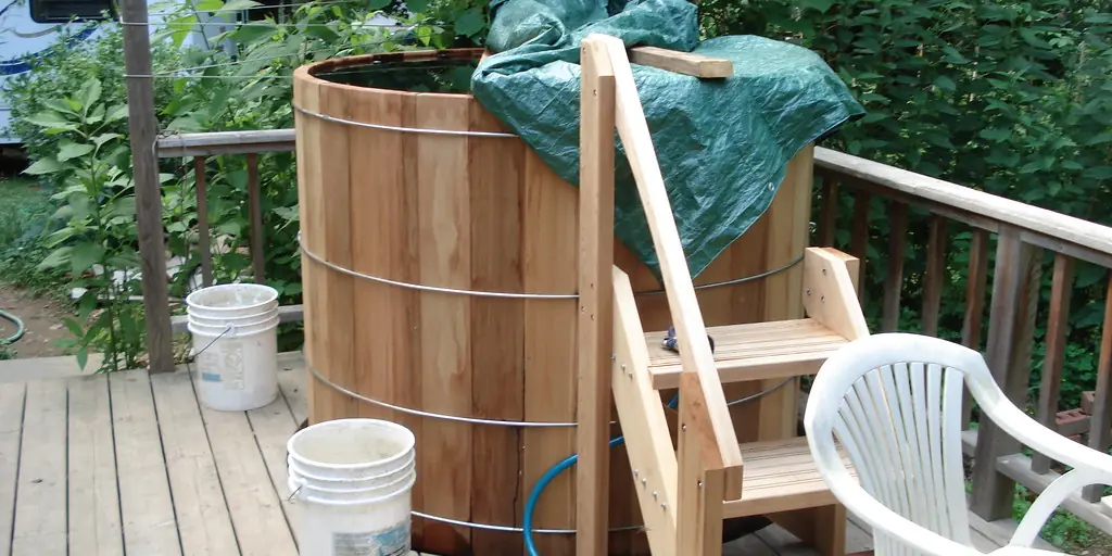 A tall cedar barrel tub bound with steel hoops standing on a garden deck, with a set of wooden steps leading up to its rim.