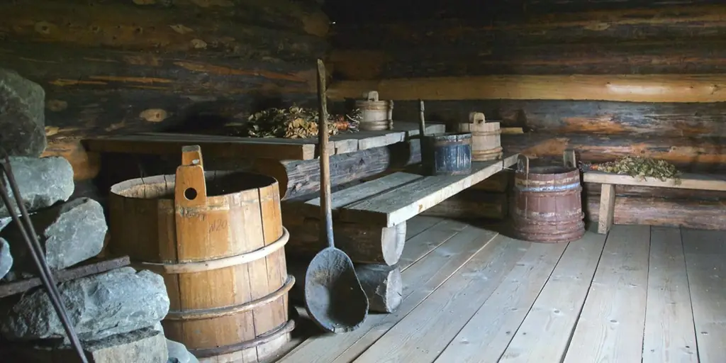 The interior of a small log bathhouse: bare timber walls and benches, a stack of stones, and a wooden bucket and dipper on the floor.